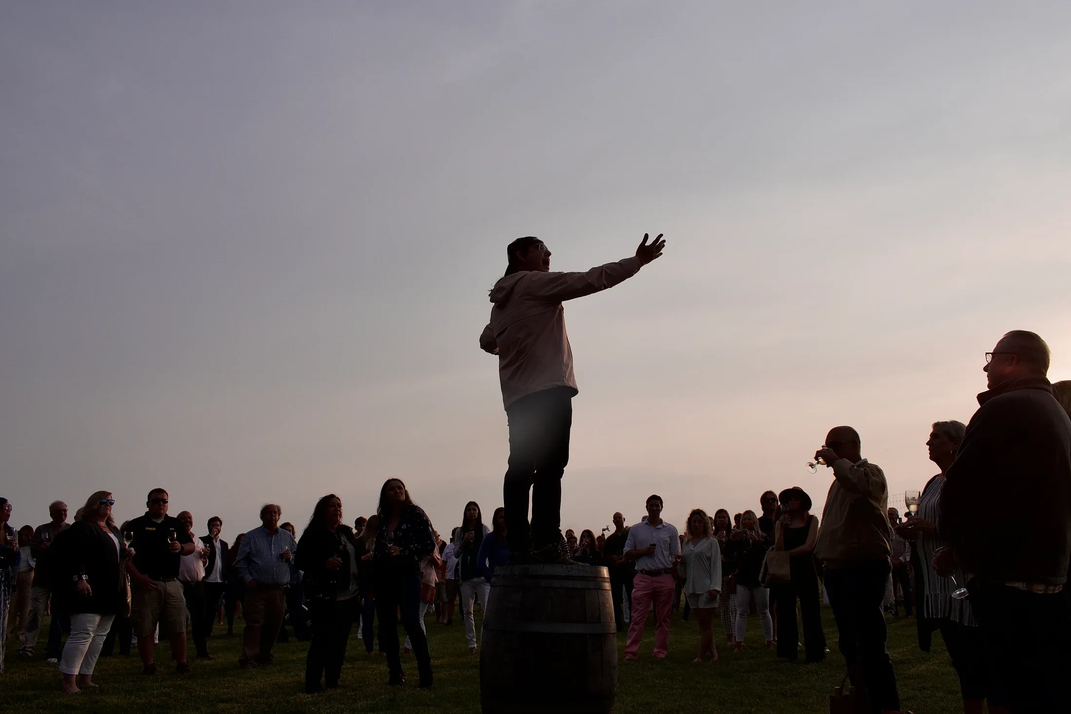 Guests gathered outside at dusk, listening to a speaker standing on a barrel