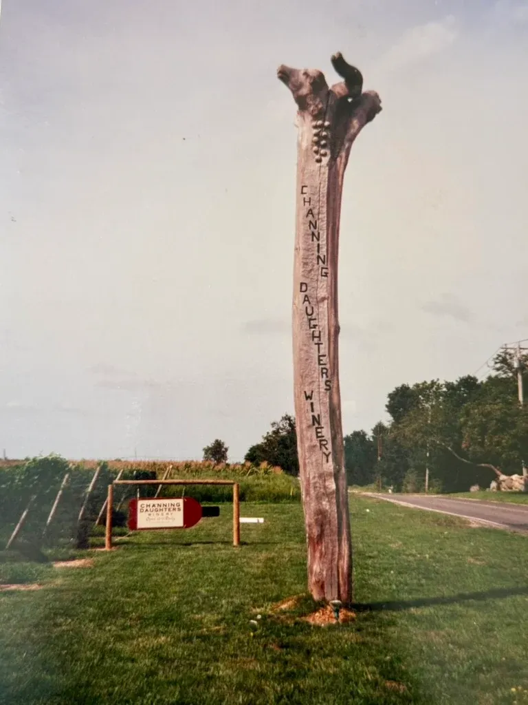 A tall carved wooden Channing Daughters Winery sign beside the vineyard entrance