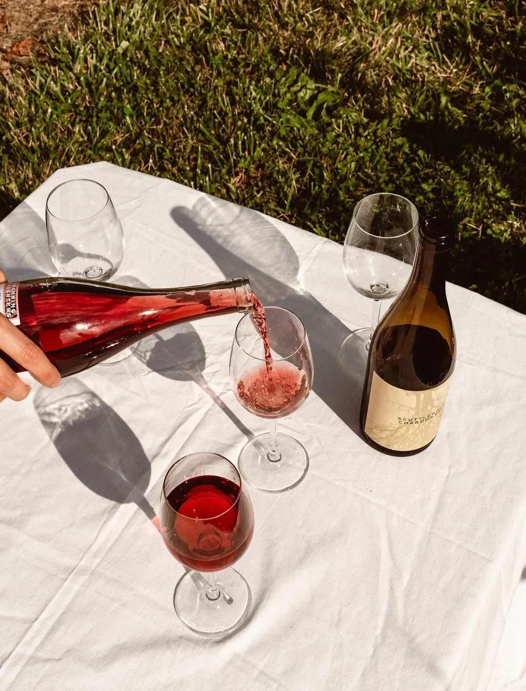 Rosé being poured into a glass beside wine bottles on a white outdoor tablecloth